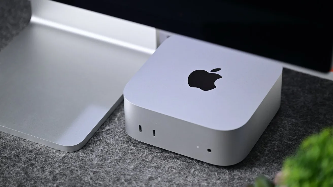 Small silver Apple Mac mini computer on a gray desktop, placed beside a metal monitor stand, with front ports and Apple logo visible, surrounded by soft, modern office lighting