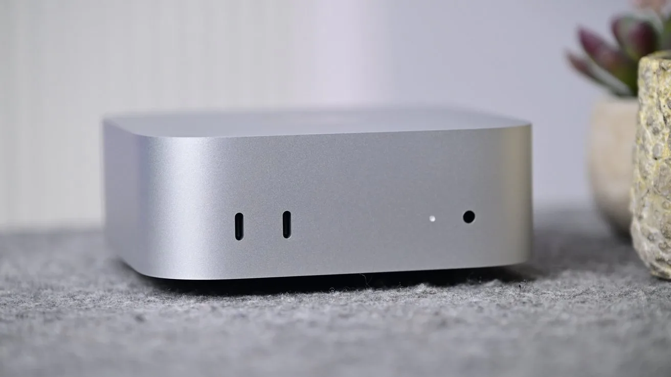 Mac mini with rounded edges on a gray surface, showing two front USB-C ports, a tiny indicator light, soft background, and a blurred plant to the right