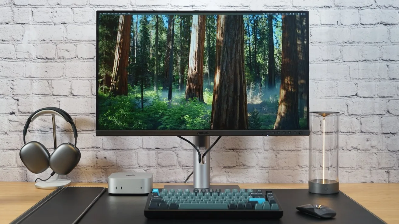 Minimal desktop setup with large monitor showing a forest scene, compact keyboard, mouse, small computer, wireless headphones on a stand, and glass cylinder, all against a white brick wall