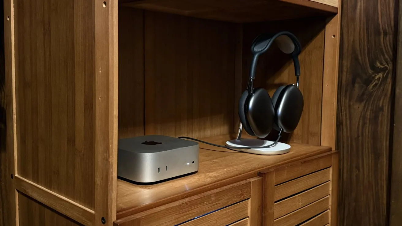 Wooden cabinet shelf holding a small silver desktop computer and large black over-ear headphones on a stand, above slatted cabinet doors in a warmly lit room