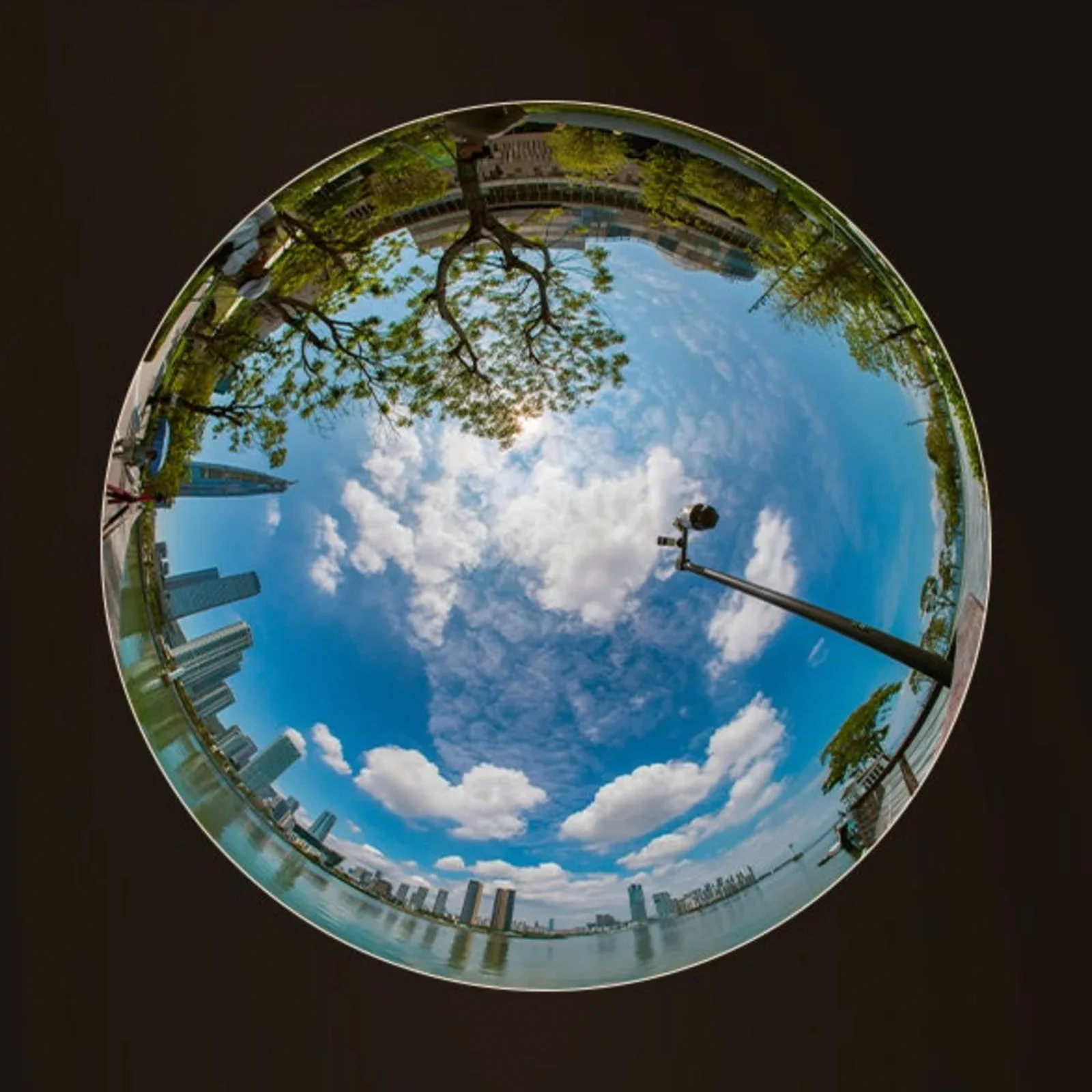 A 360-degree fisheye view of a city skyline along a waterfront under a bright blue sky with scattered clouds.
