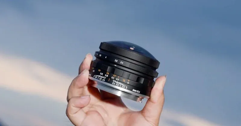A hand holds a black fisheye camera lens against a blurred blue sky background.