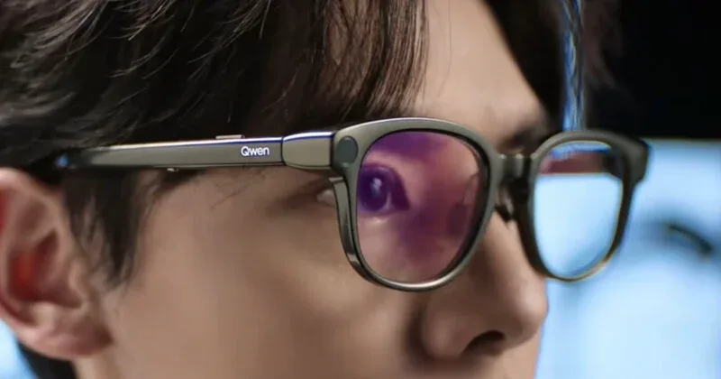 A close-up side view of a person wearing dark-rimmed glasses with the word "Qwen" printed on the temple.
